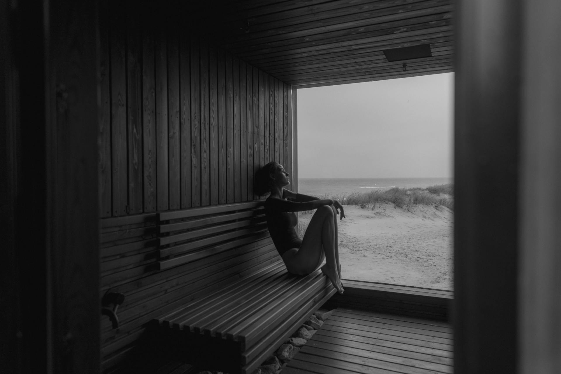 A woman leaning back bathing in a sauna next to a lake on the sand.