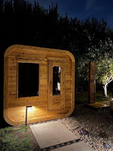 A Henki Cube Sauna at night with a light out front and an outdoor shower next to it.