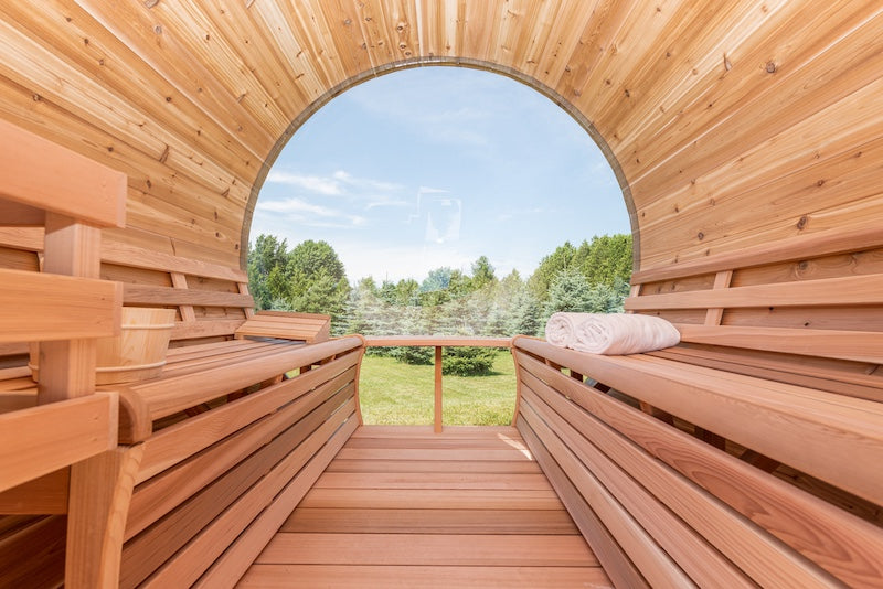 The inside of a Henki Custom Barrel Sauna with clear cedar benches and a large dome window overlooking a green landscape.