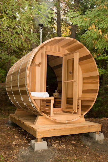 A Henki Custom Barrel Sauna on a small porch in the BC forest with smoke coming out of the chimney. 