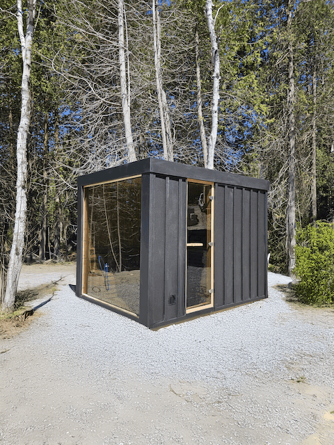 A Pure Cube Neptune Sauna with black metal siding on a gravel patch.