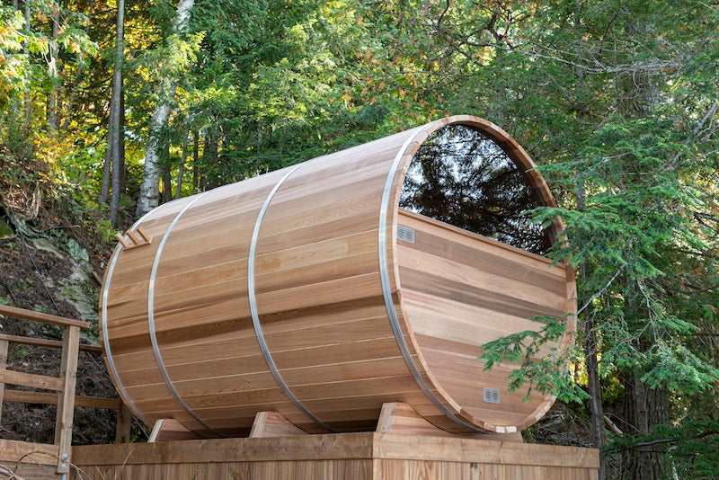 The back and side of a Henki Panoramic Barrel Sauna on a deck in the woods in Muskoka.
