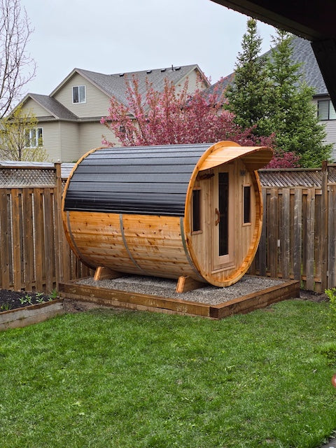 A Henki Window Barrel Sauna with a black metal roof and a cedar cove roof overhang.