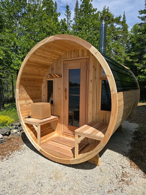 A Henki Window Barrel Sauna with a black metal roof a box of rocks on the porch bench.