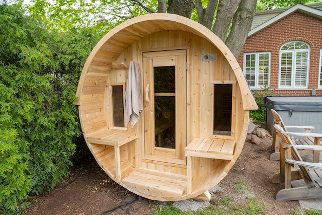 A Henki Window Barrel Sauna in the backyard of a brick house next to Muskoka chairs and a hot tub.